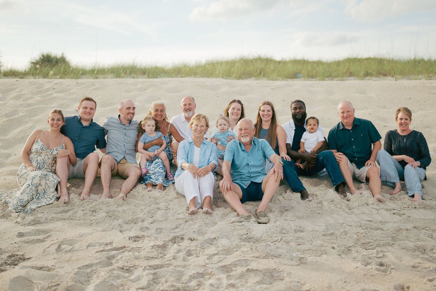 Large family group portrait on sandy beach, smiling and sitting together in coordinated blue and white outfits.