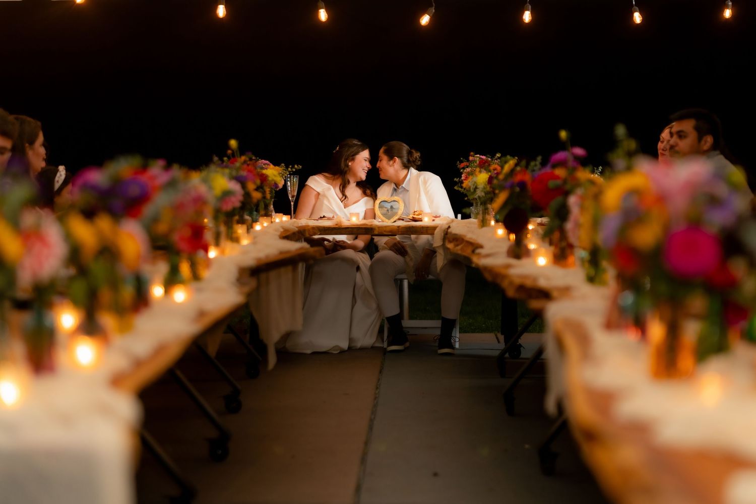 Couple shares intimate moment at candlelit wedding reception table adorned with colorful floral centerpieces.