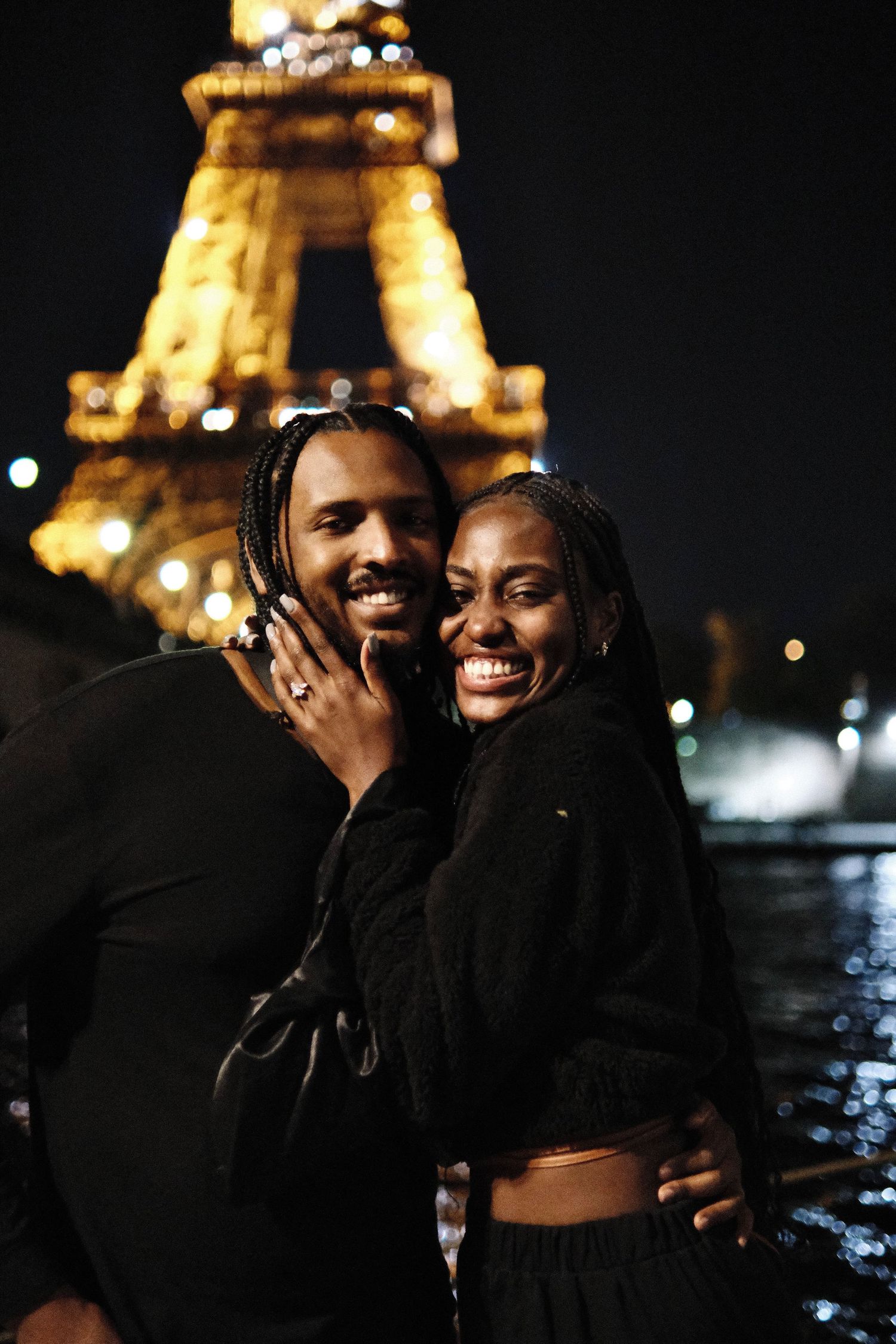 A smiling couple embraces at night in front of the illuminated Eiffel Tower in Paris, France.