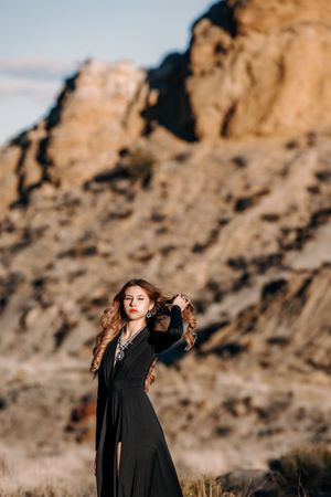 Woman in black dress poses gracefully before rugged desert rock formations at golden hour.