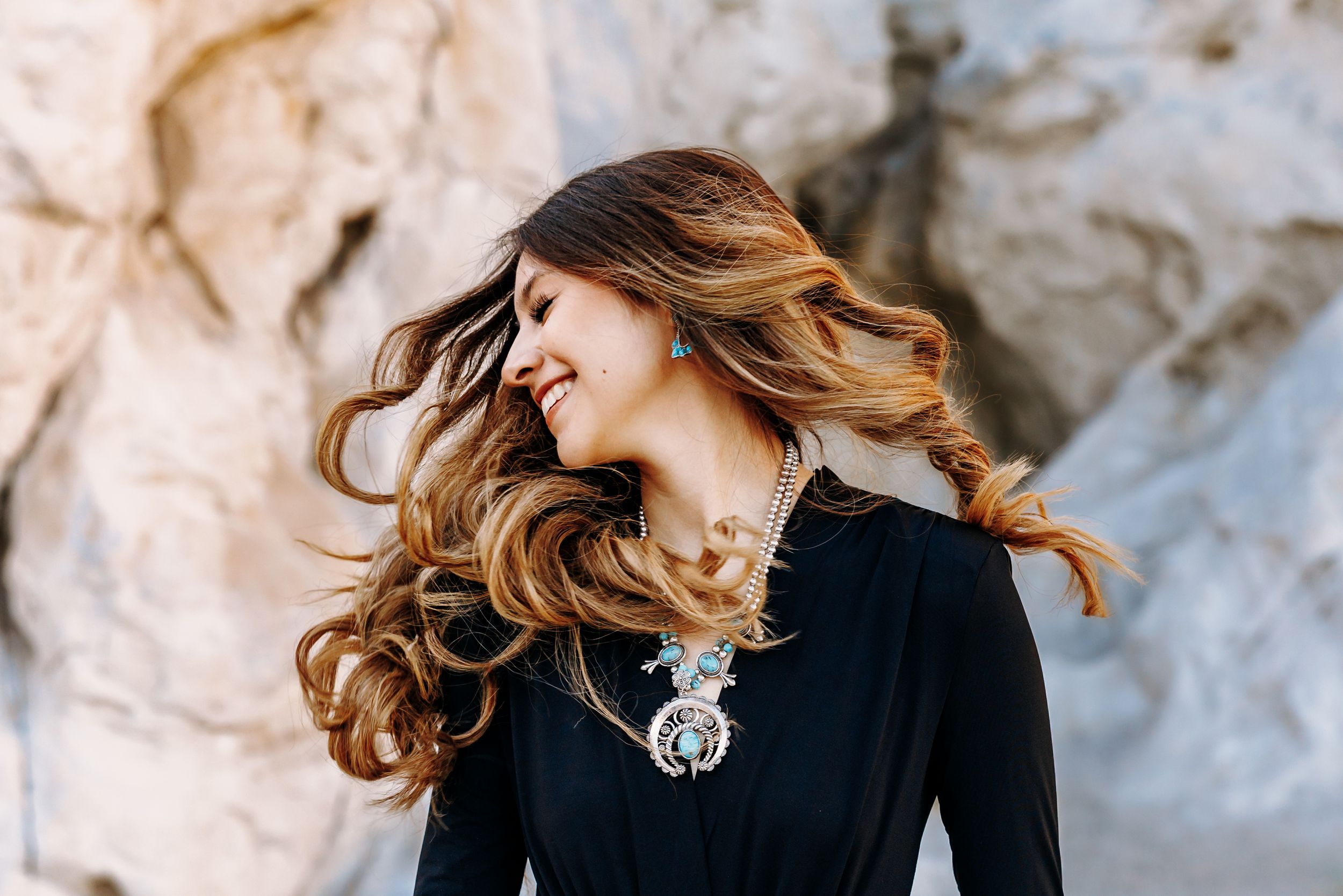 Woman with flowing ombre hair smiling, wearing turquoise jewelry and black dress against rocky background.