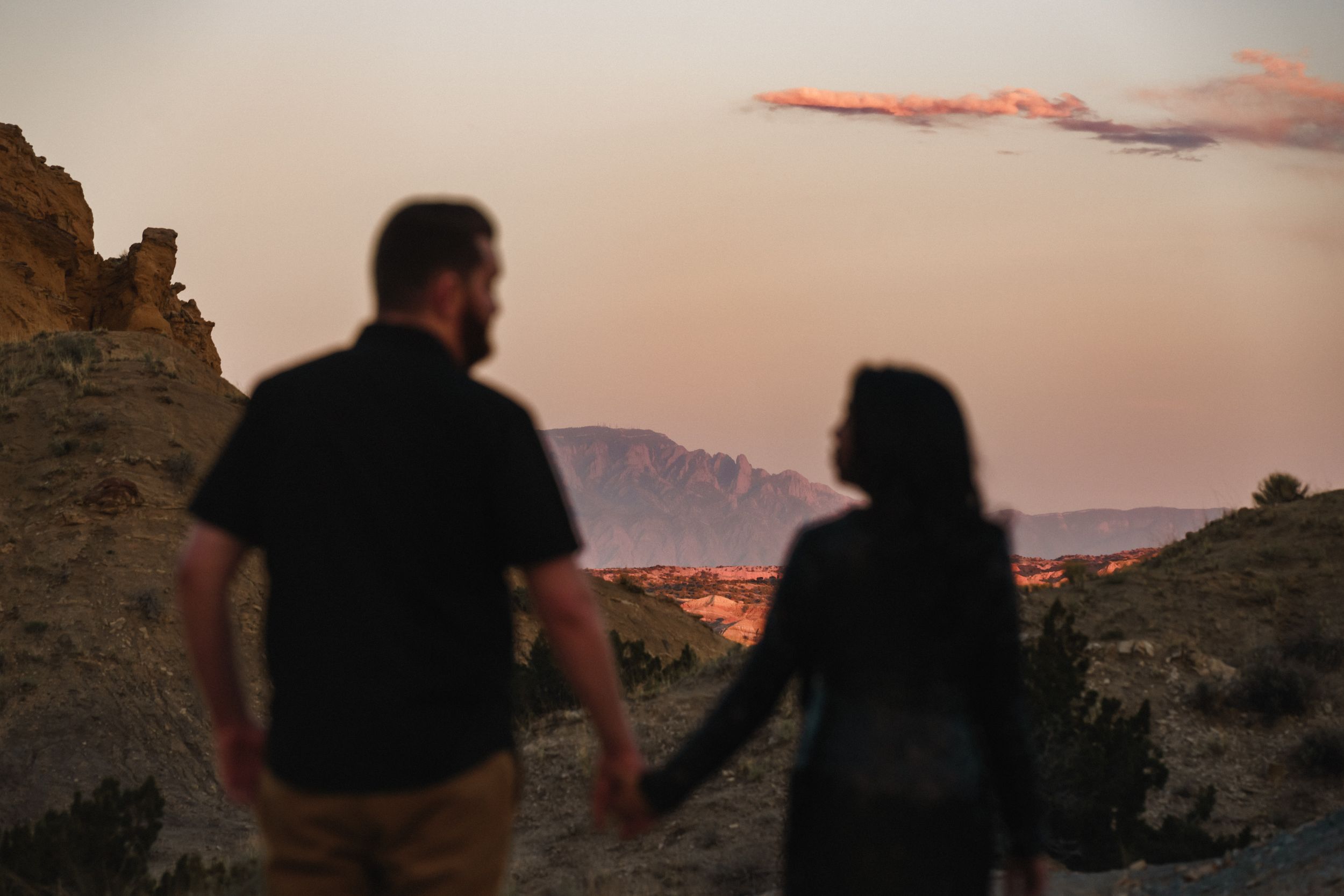 Couple holding hands at sunset overlooking a desert landscape with mountains and a glowing pink sky.