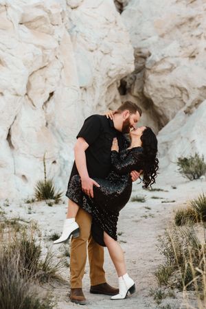 Couple sharing a romantic dip kiss in front of white rock formations, woman in sequin dress and white boots.