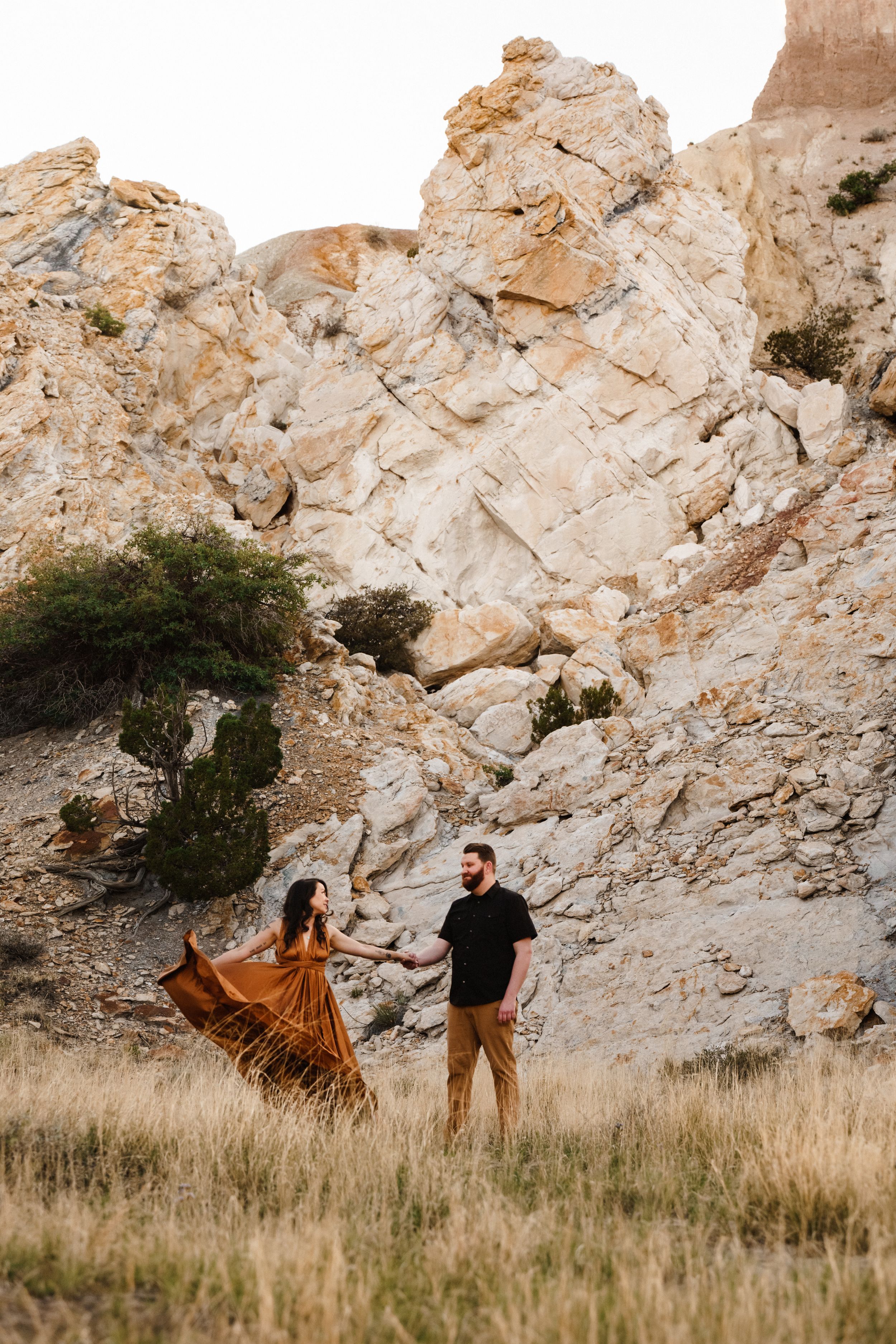 Couple holding hands in a golden field with dramatic rocky cliffs behind them, woman in flowing rust dress.