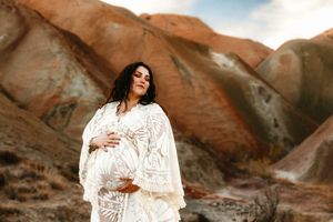Pregnant woman in white lace dress cradling belly against colorful painted hills landscape.