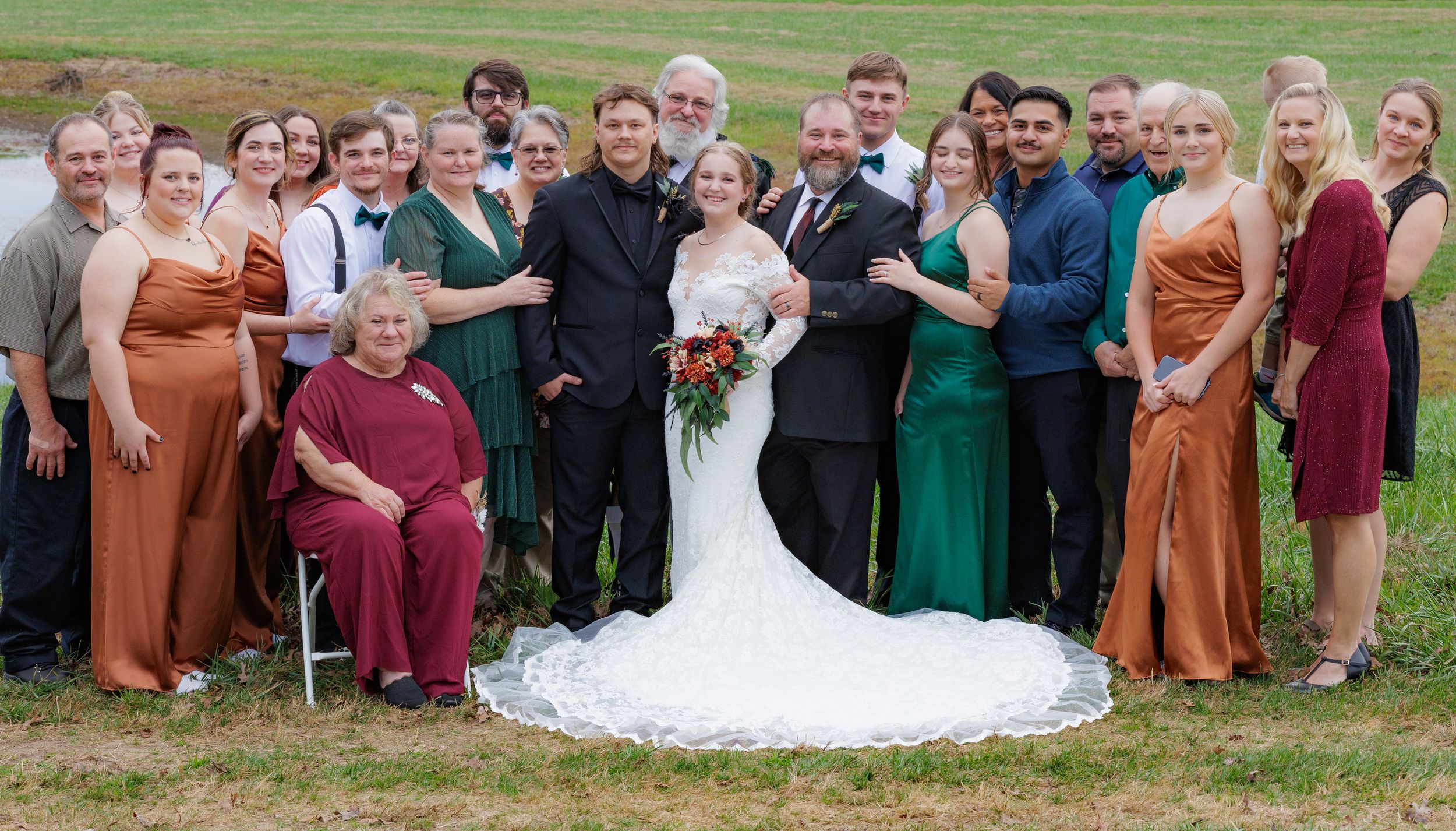 A large wedding party poses together outdoors wearing formal attire in autumn colors and a white wedding gown.