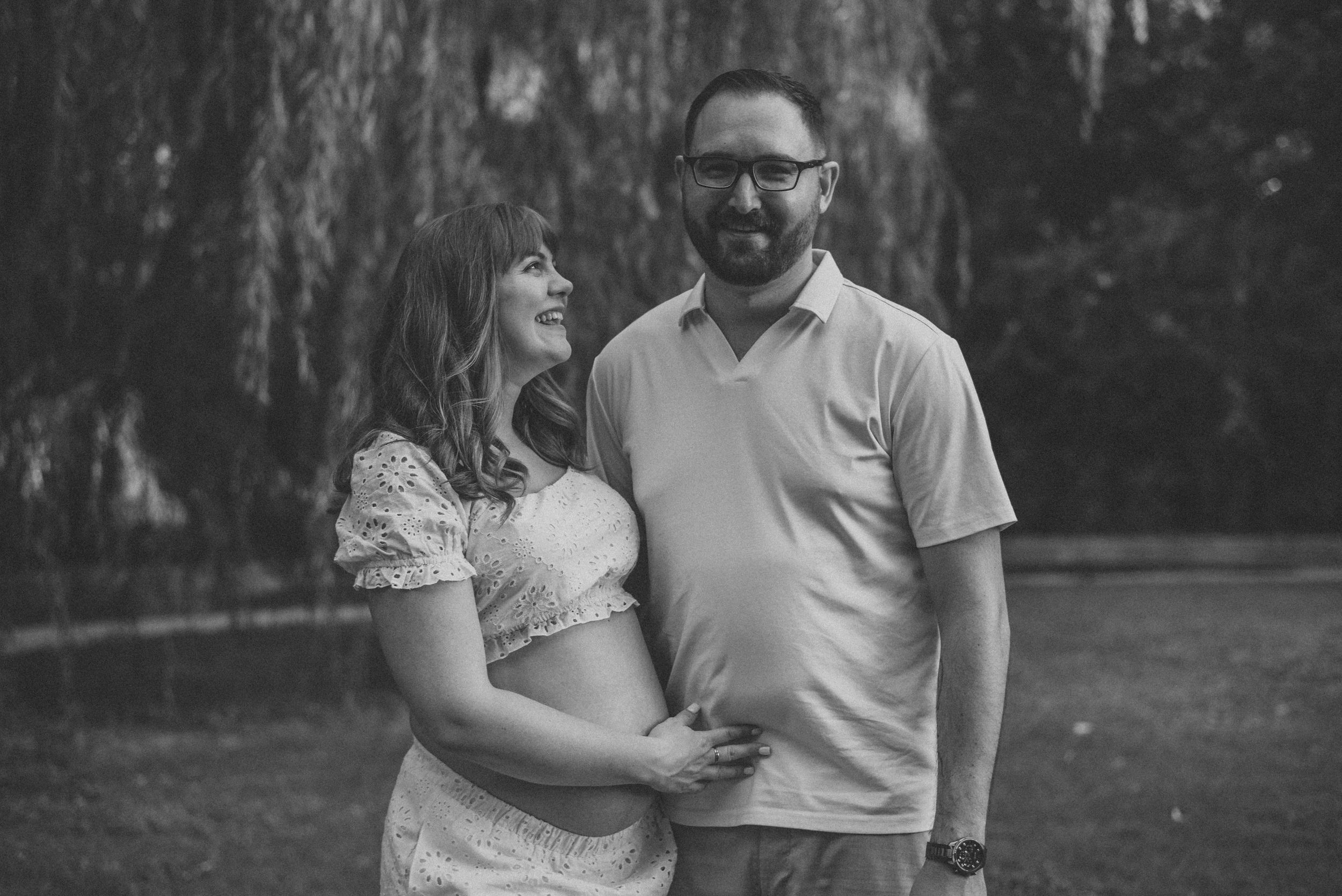 Black and white photo of a smiling couple embracing outdoors near a weeping willow tree.