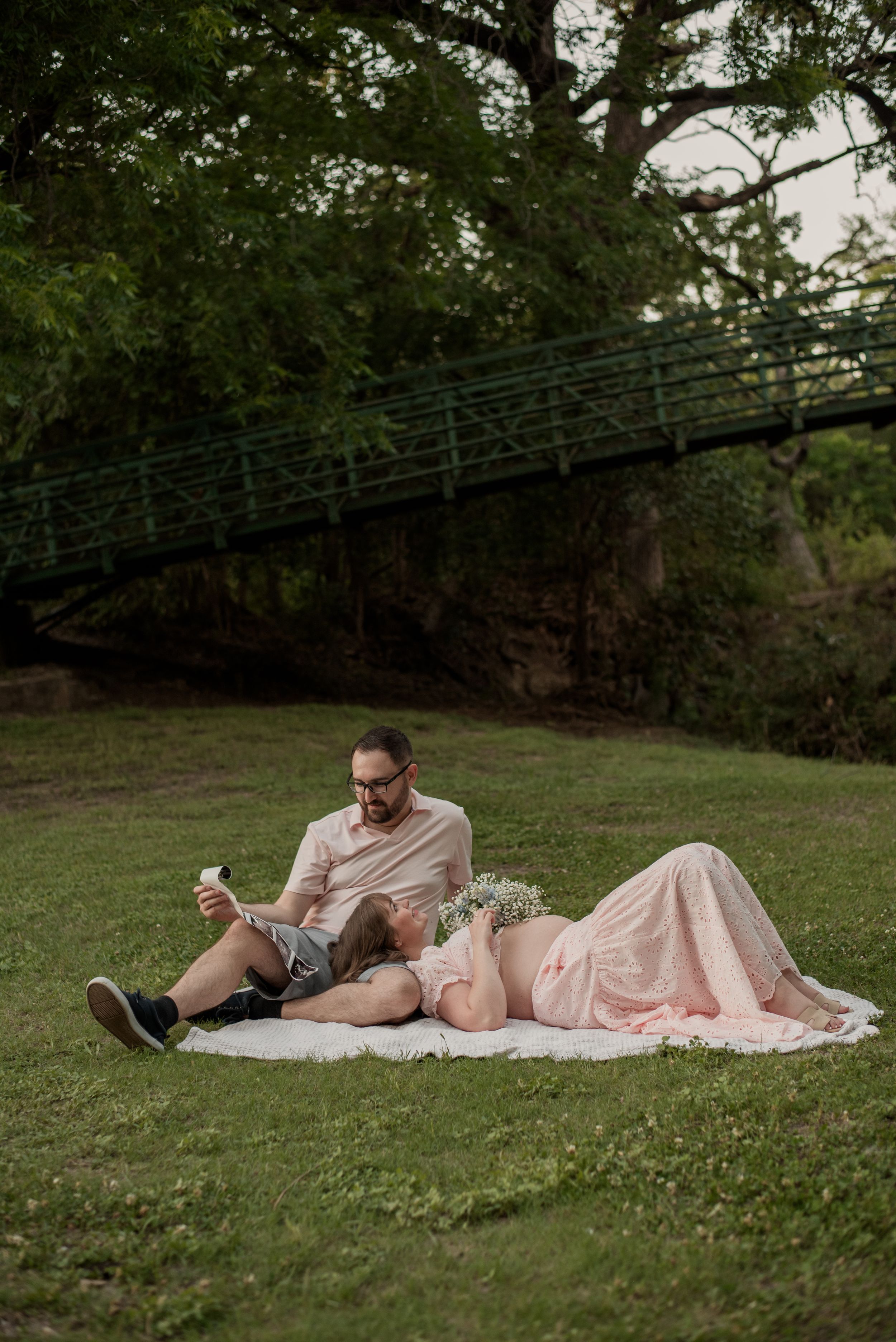 Pregnant couple relaxing on a blanket in a park, man holding phone while woman in pink dress lies with flowers.