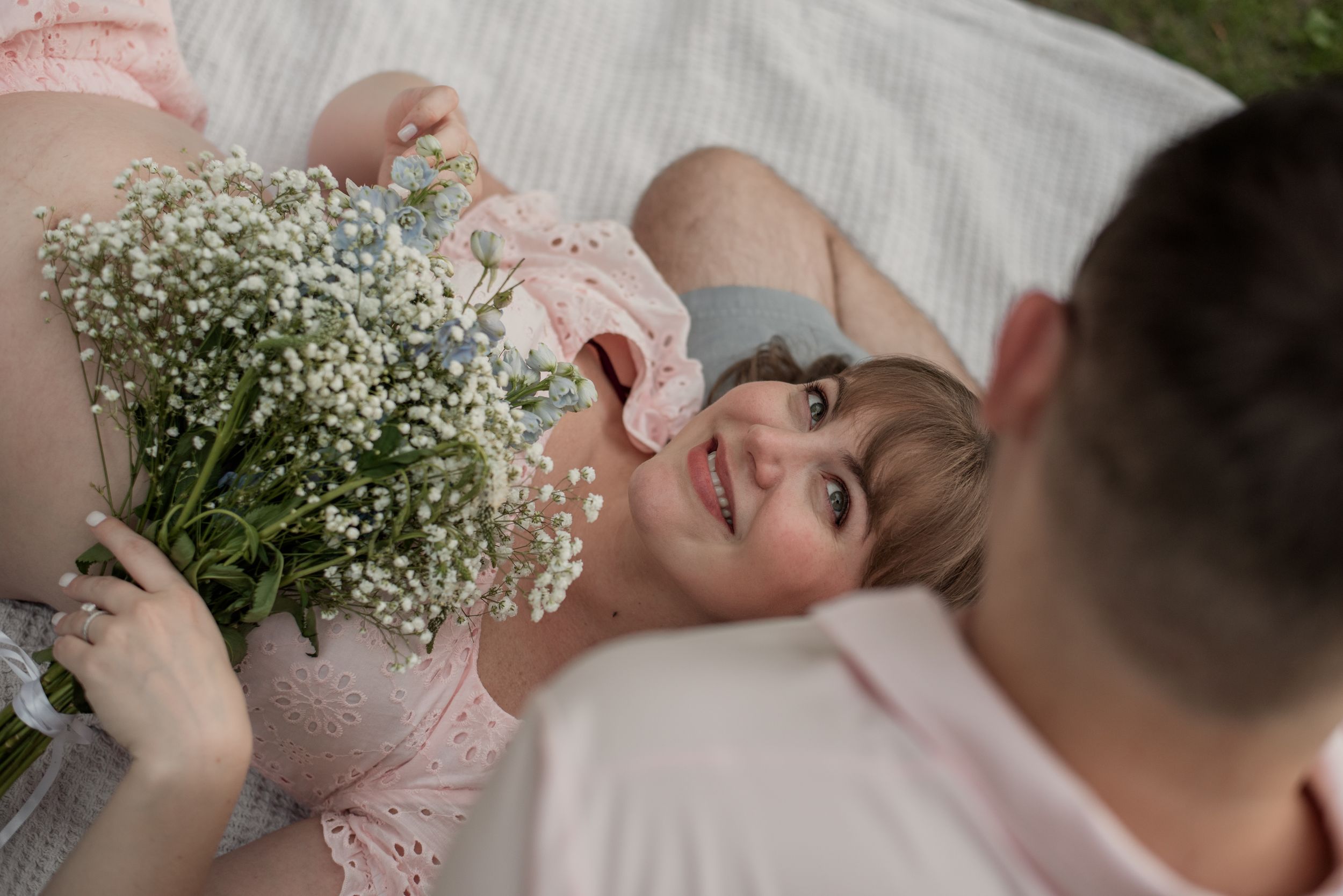 Pregnant woman smiling while lying on blanket holding white wildflowers bouquet with partner beside her outdoors.