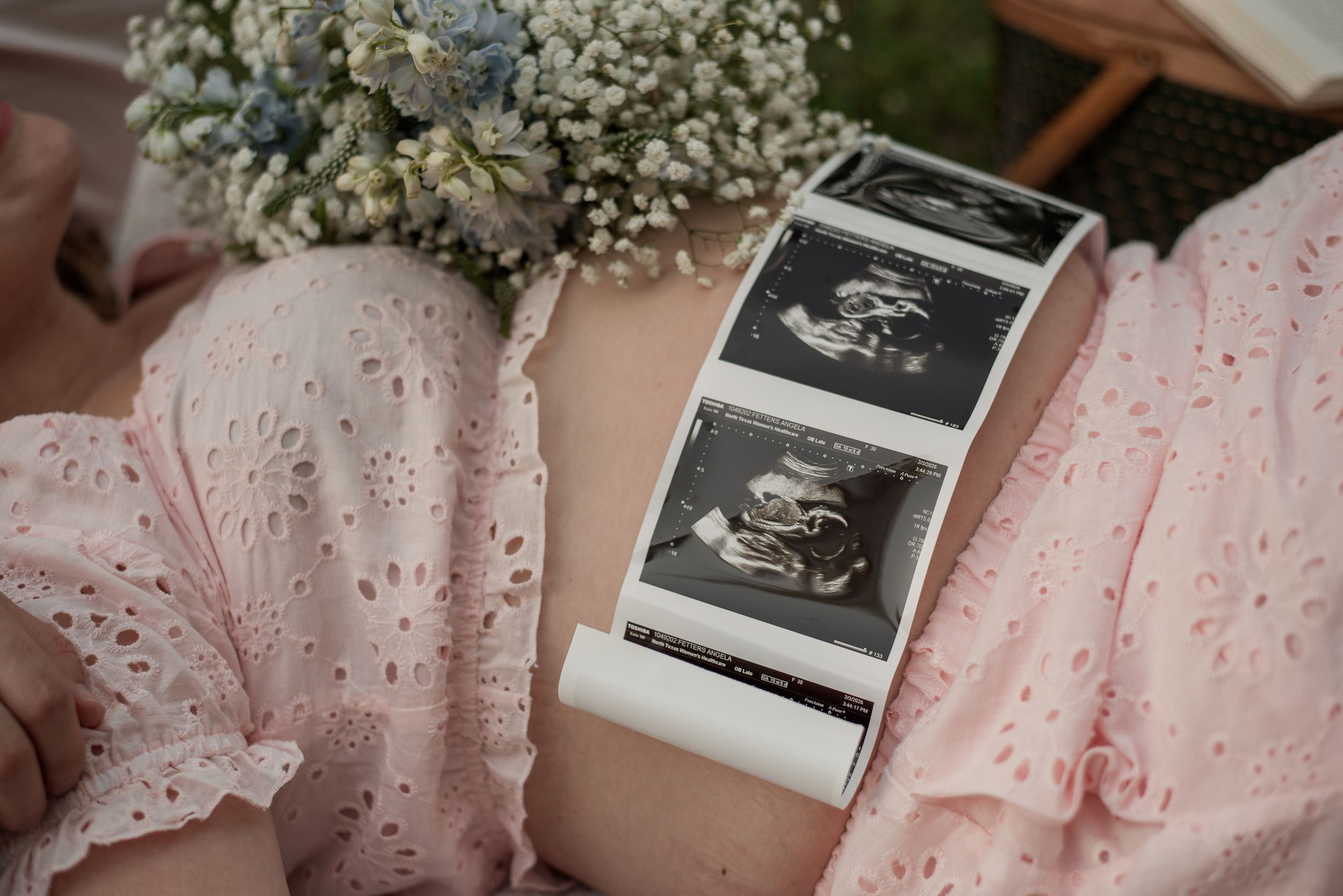 Pregnant woman in pink dress holding ultrasound scan photos with blue and white flowers in background.