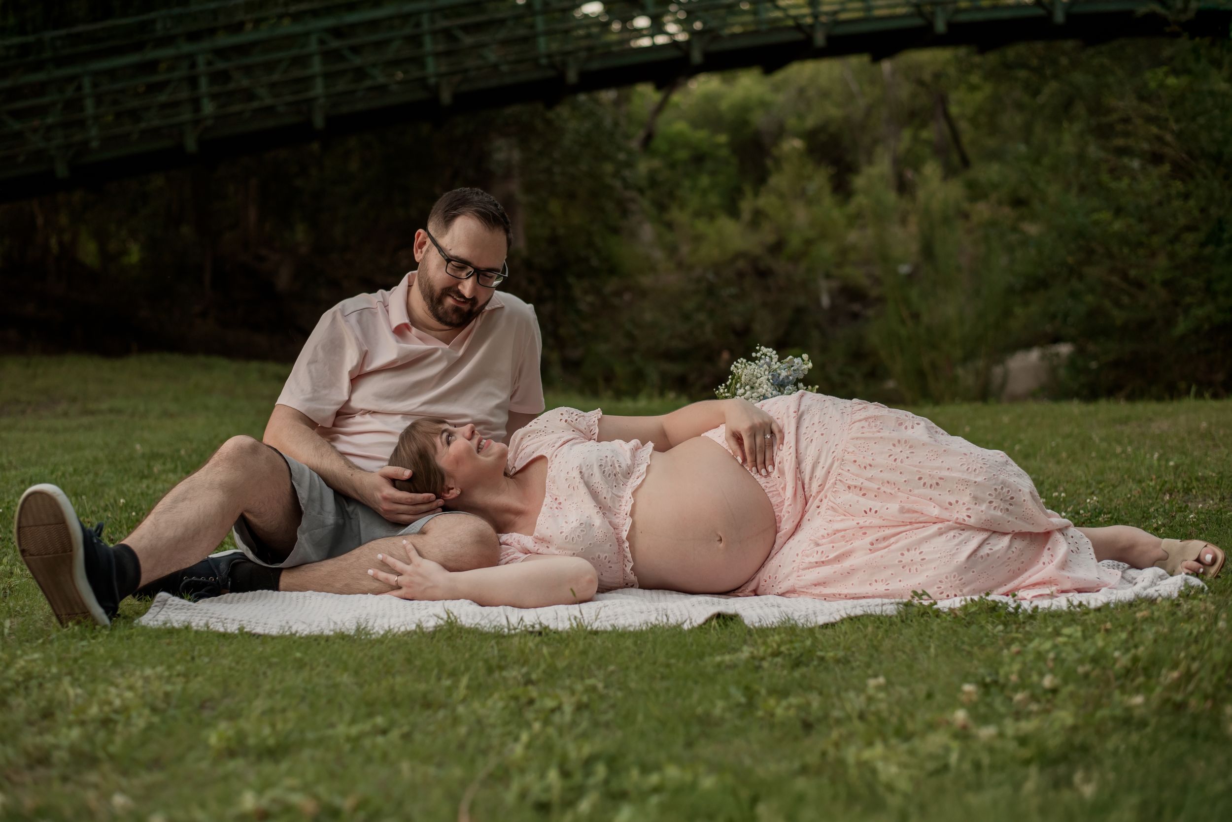 Pregnant couple relaxing on a blanket in a park, woman in pink dress lying down, man sitting beside her reading.
