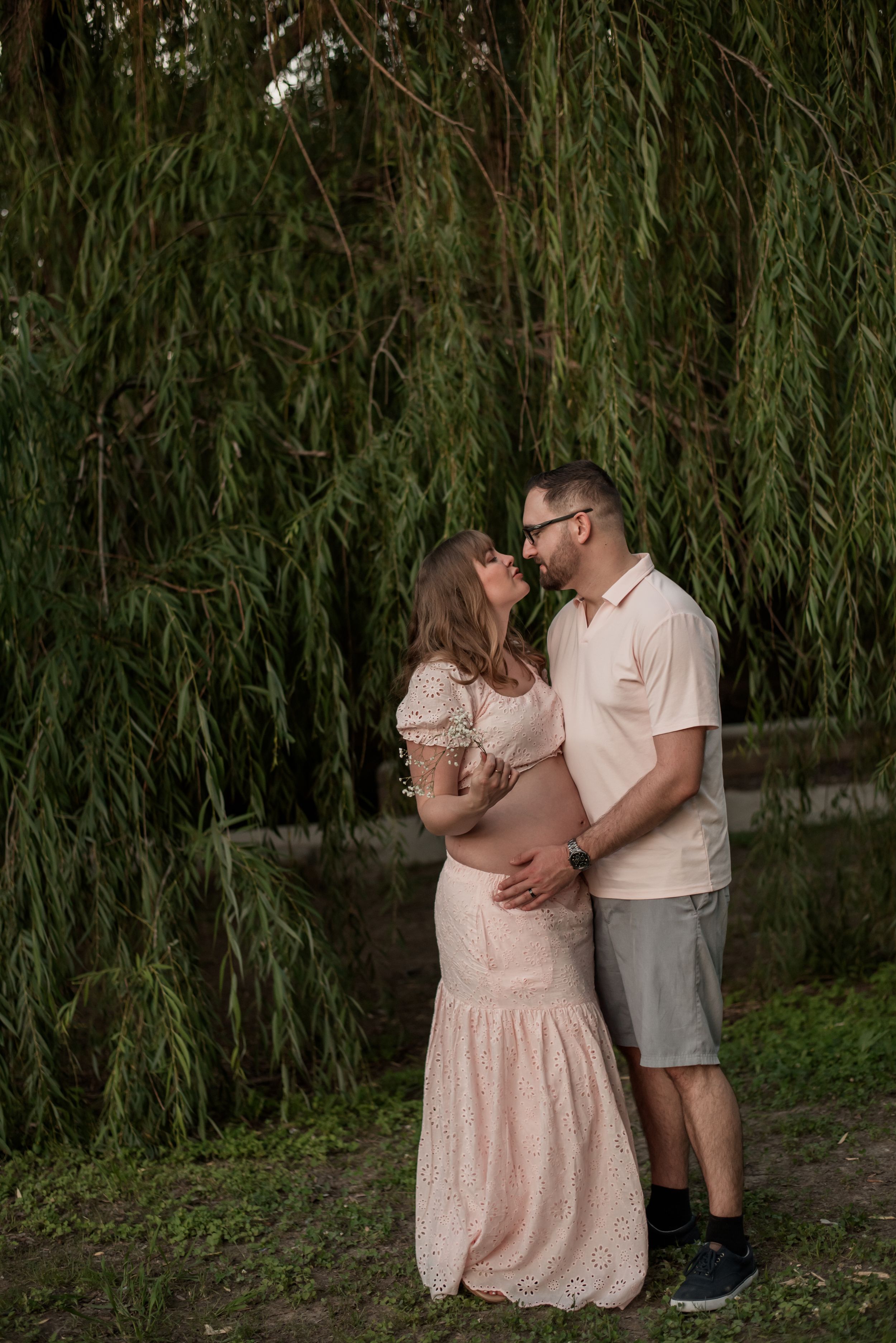 Couple sharing a tender moment outdoors under a weeping willow tree, with the woman wearing a pink lace maternity dress.