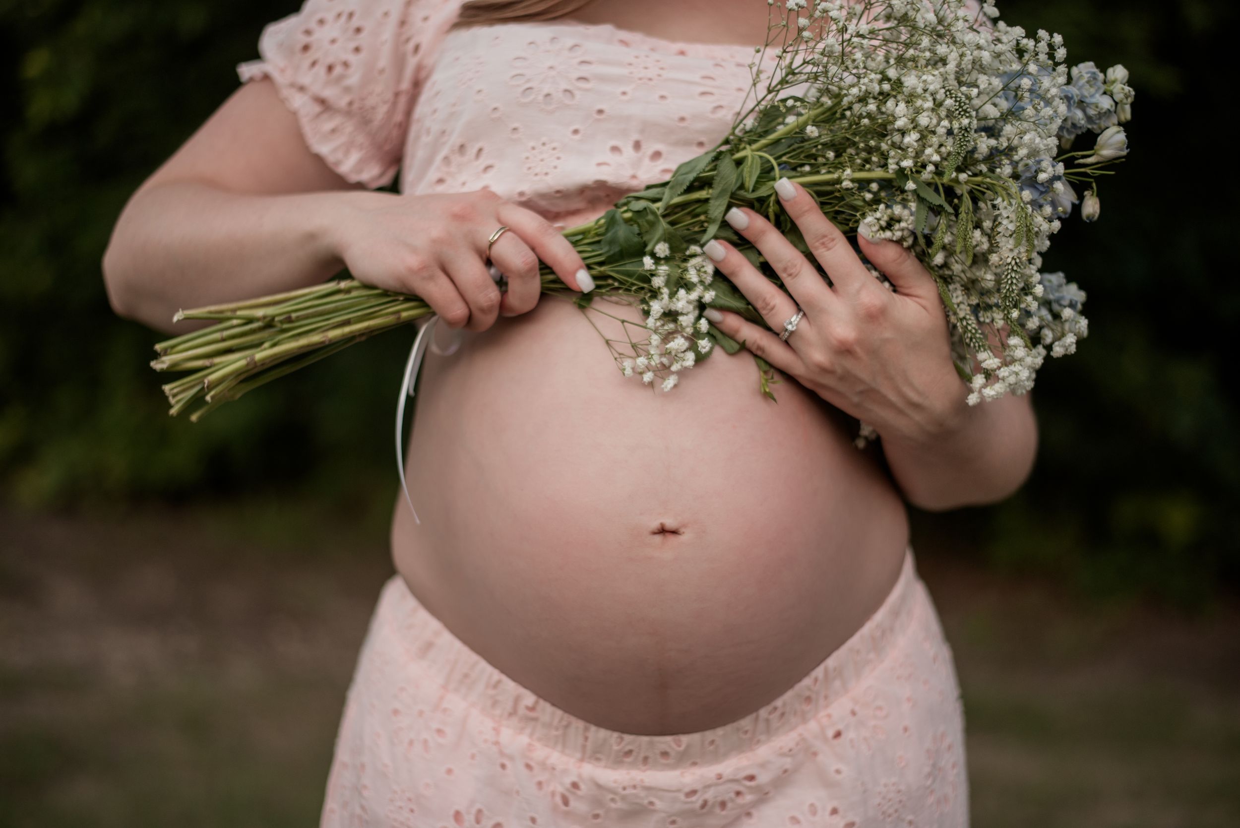 Pregnant woman in pink lace dress holding wildflower bouquet, showcasing baby bump outdoors.