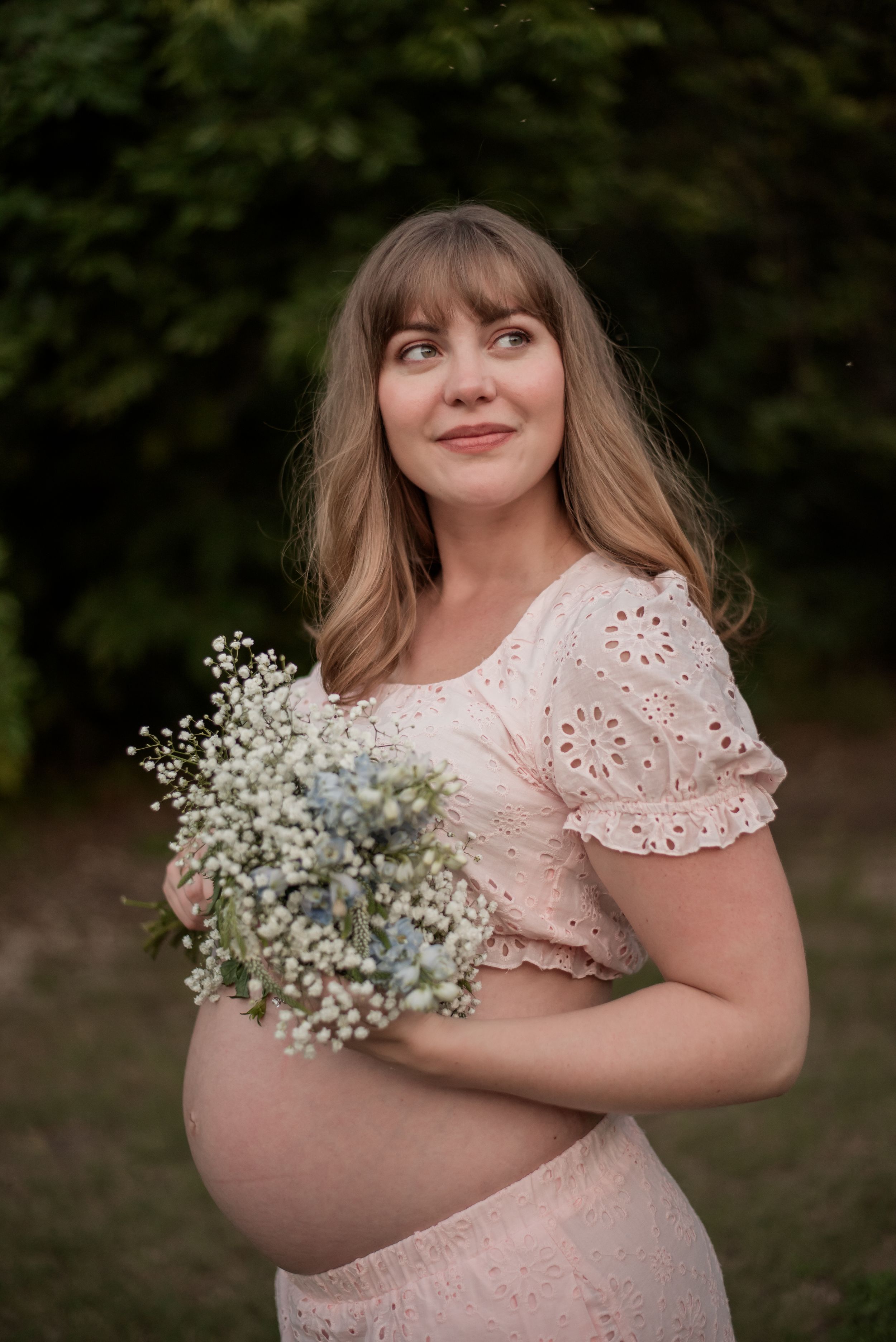 Pregnant woman in pink eyelet dress holding baby's breath bouquet outdoors, smiling softly.