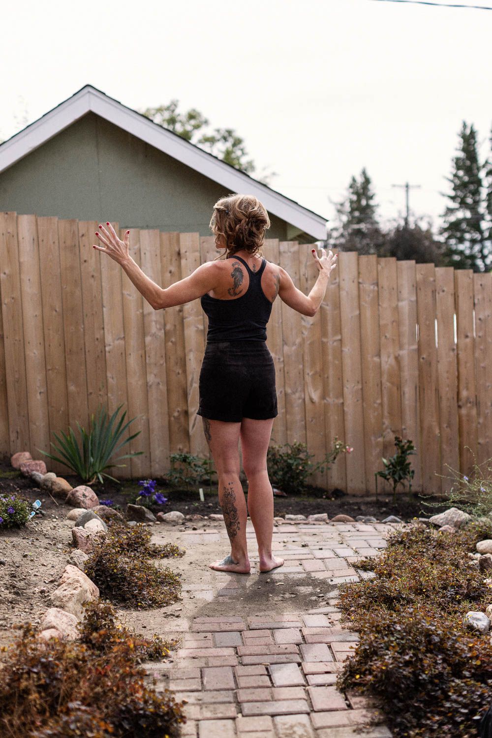 Woman in black outfit standing on brick path in backyard, arms raised, facing wooden fence.