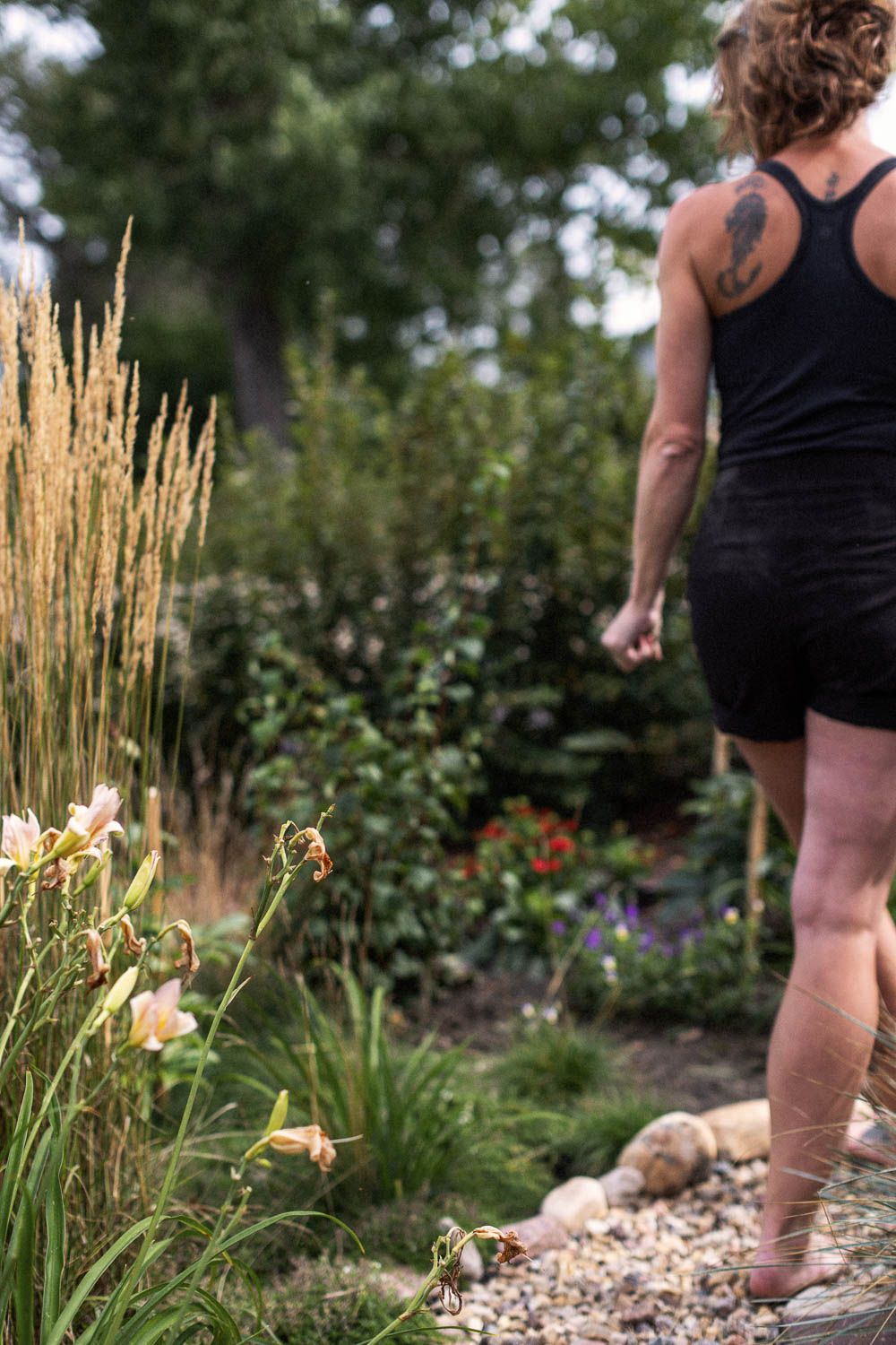 Woman with tattoo in black tank top and shorts walking along a garden path with ornamental grasses and flowers.