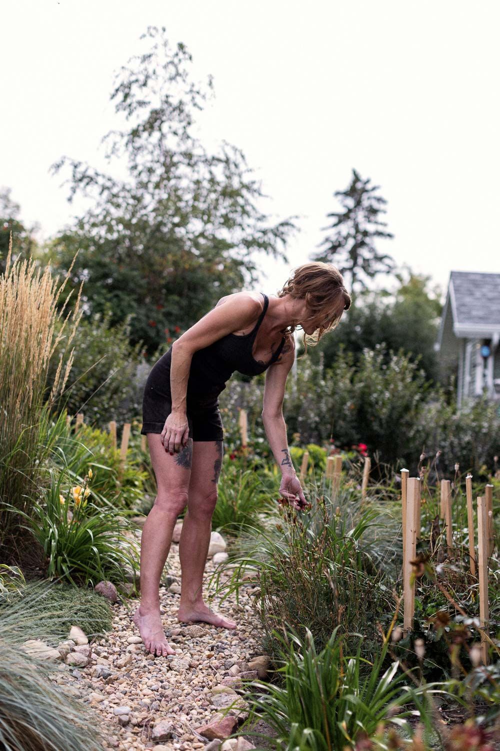 Woman in black shorts tending to plants barefoot on a gravel garden path surrounded by lush greenery.