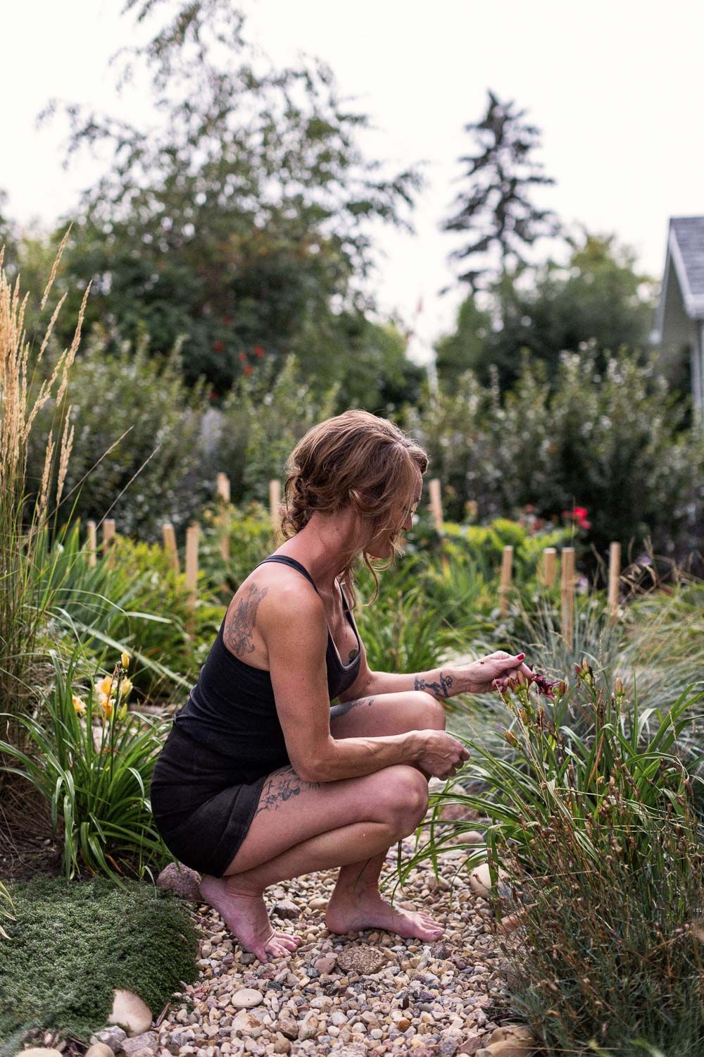 A barefoot woman with tattoos crouches in a lush garden, tending to plants along a gravel path.