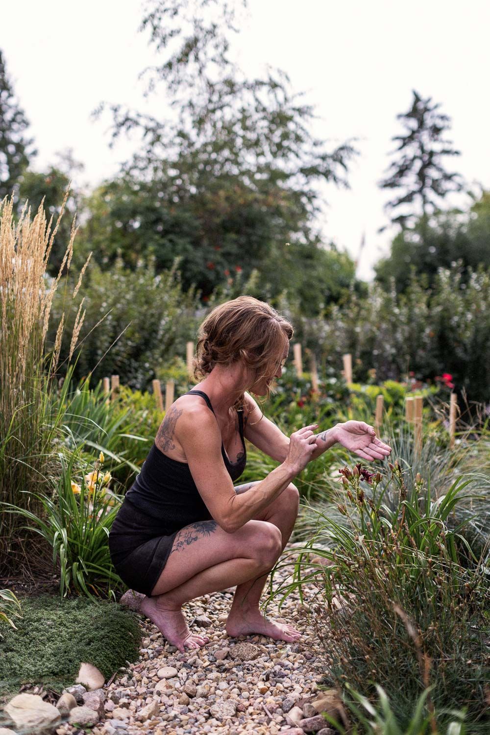 Woman crouching barefoot in a lush garden, examining plants along a gravel path surrounded by greenery.