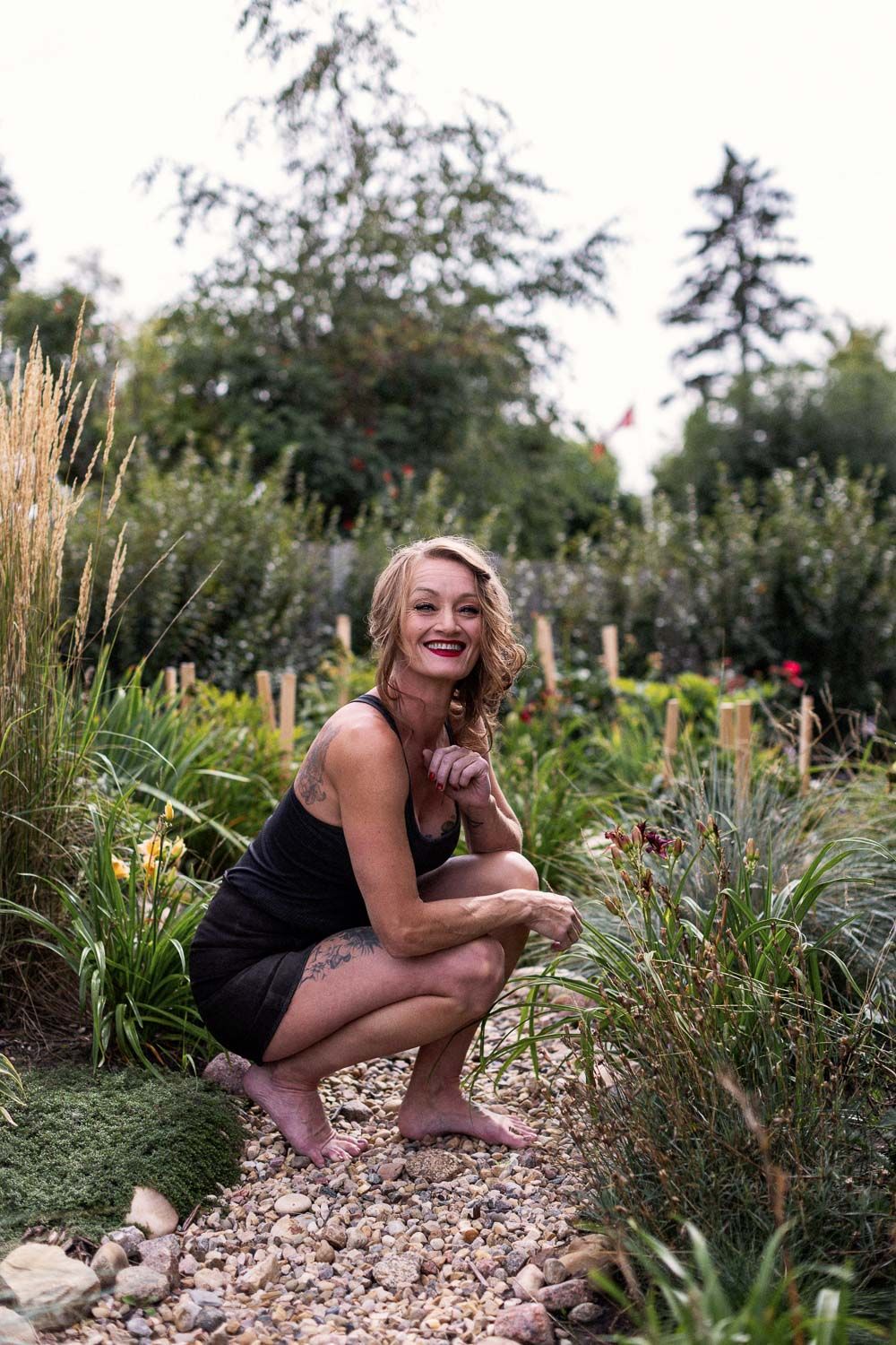Smiling blonde woman crouching barefoot in a lush garden, wearing a black outfit with visible tattoos.