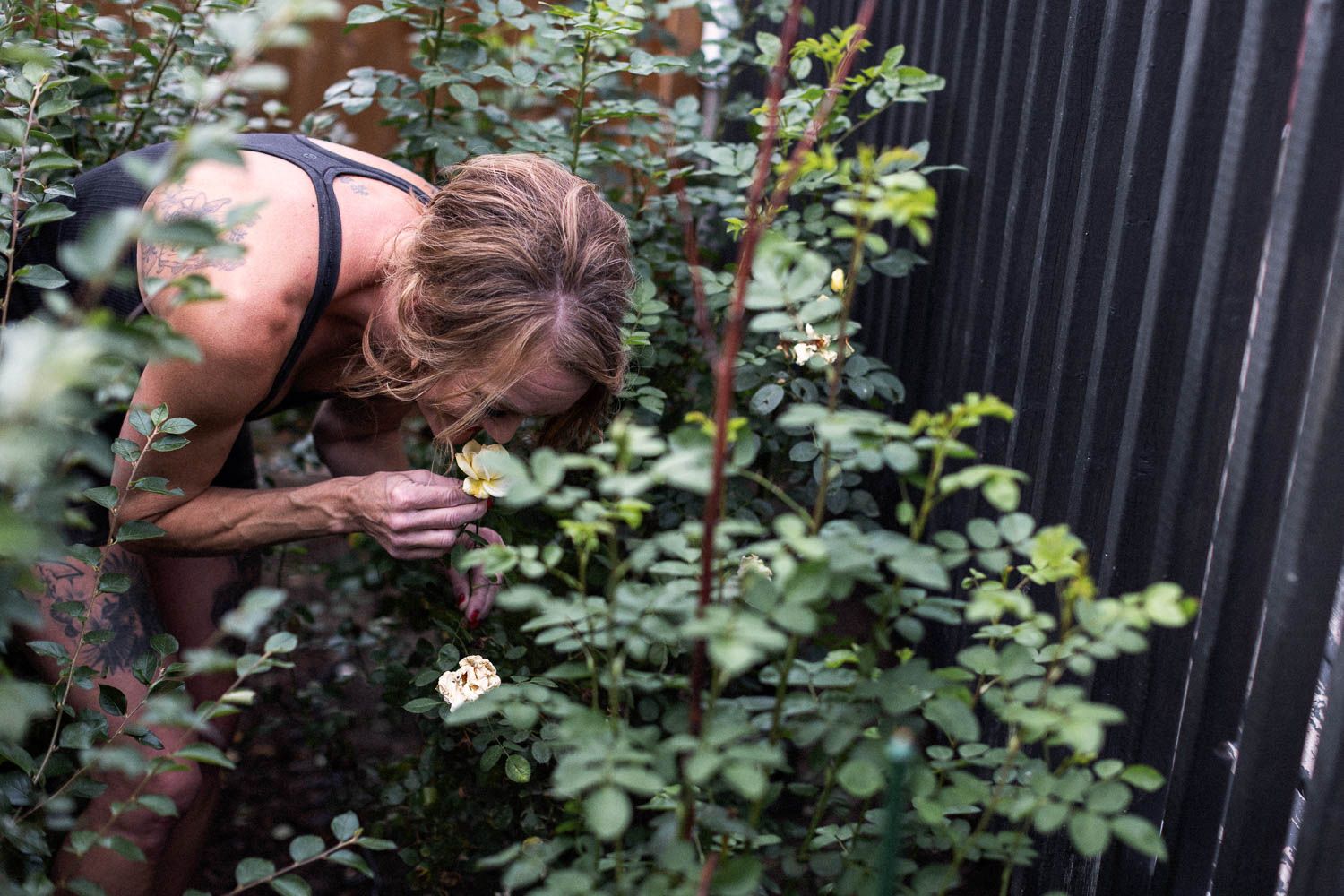 Woman leaning over green garden shrubs smelling a small white flower near a dark wooden fence.