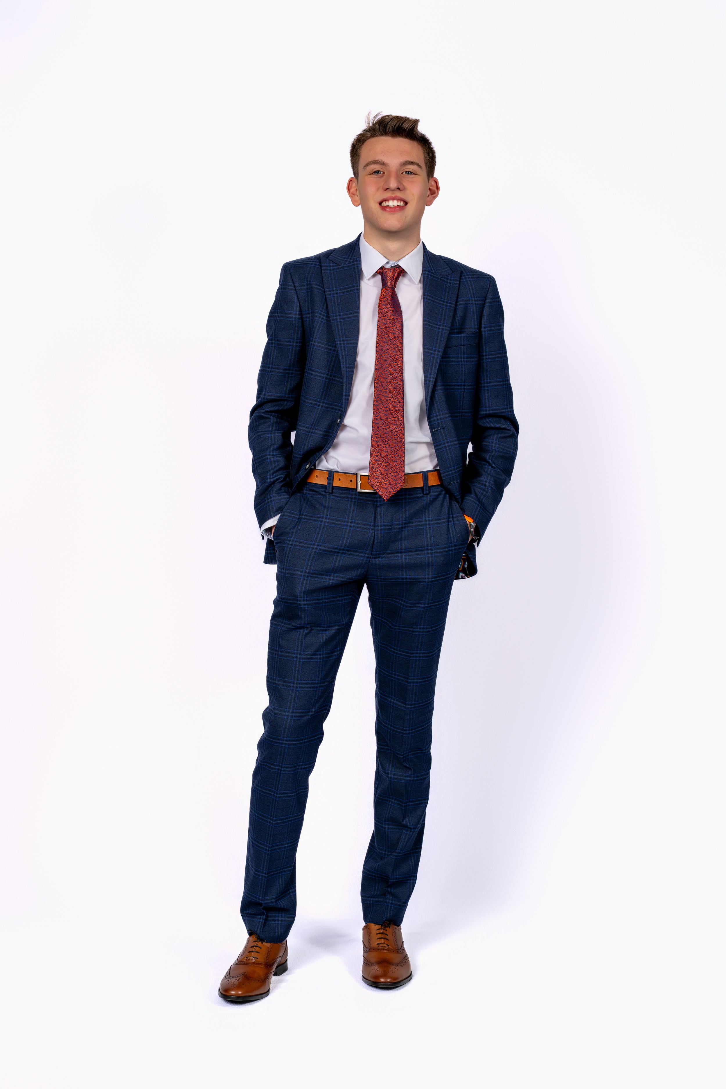 Young man in navy blue suit, red tie, and brown dress shoes posing confidently with hands in pockets.
