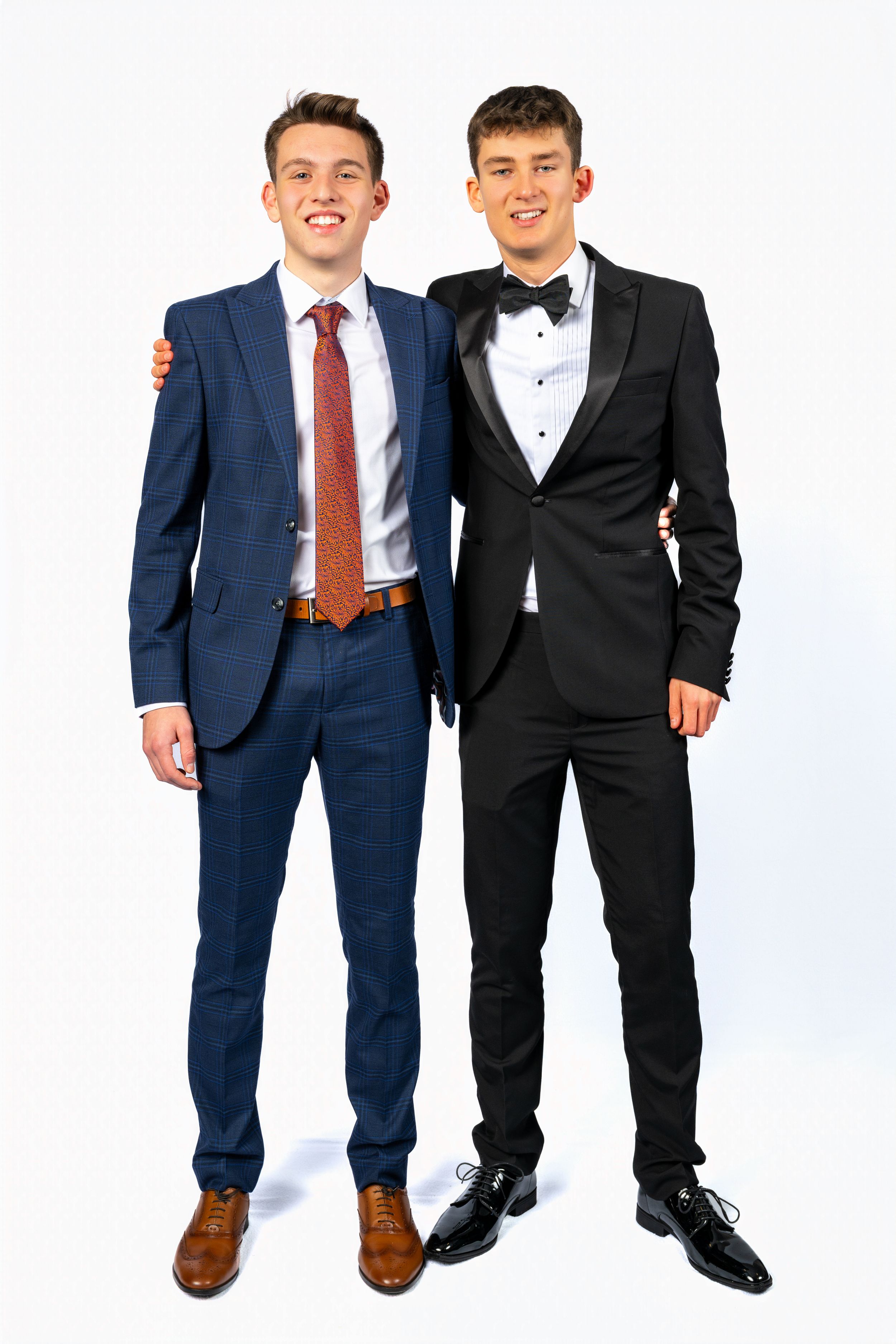 Two young men in formal suits smiling together, one in navy blue with red tie, other in black tuxedo.