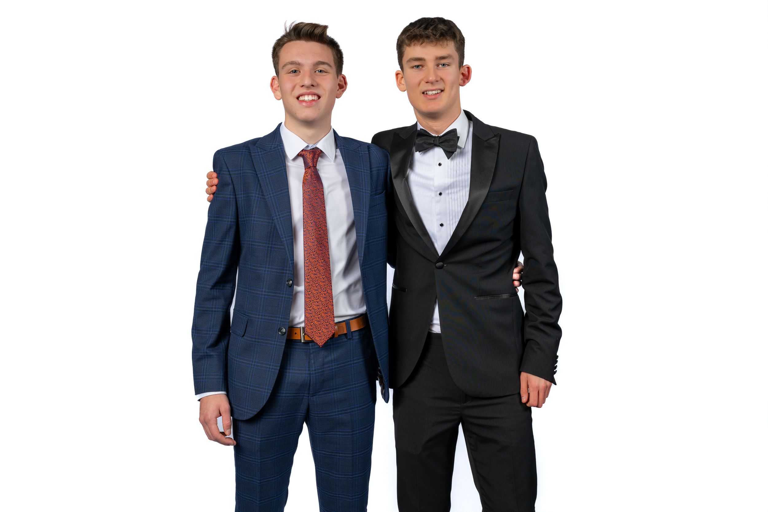 Two young men in formal suits smiling together, one in a navy blue suit with red tie, the other in a black tuxedo.