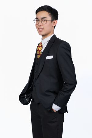 Young Asian businessman in black three-piece suit, polka dot tie, and glasses smiling against white background.