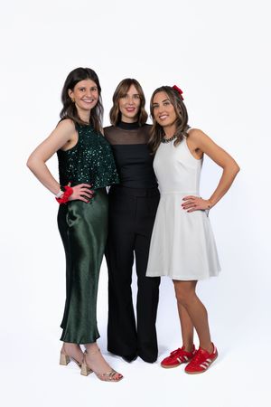 Three women posing together in festive outfits: green sequin top, black jumpsuit, and white dress with red sneakers.