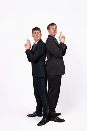 Two young men in suits stand back-to-back pointing finger guns in a studio setting.