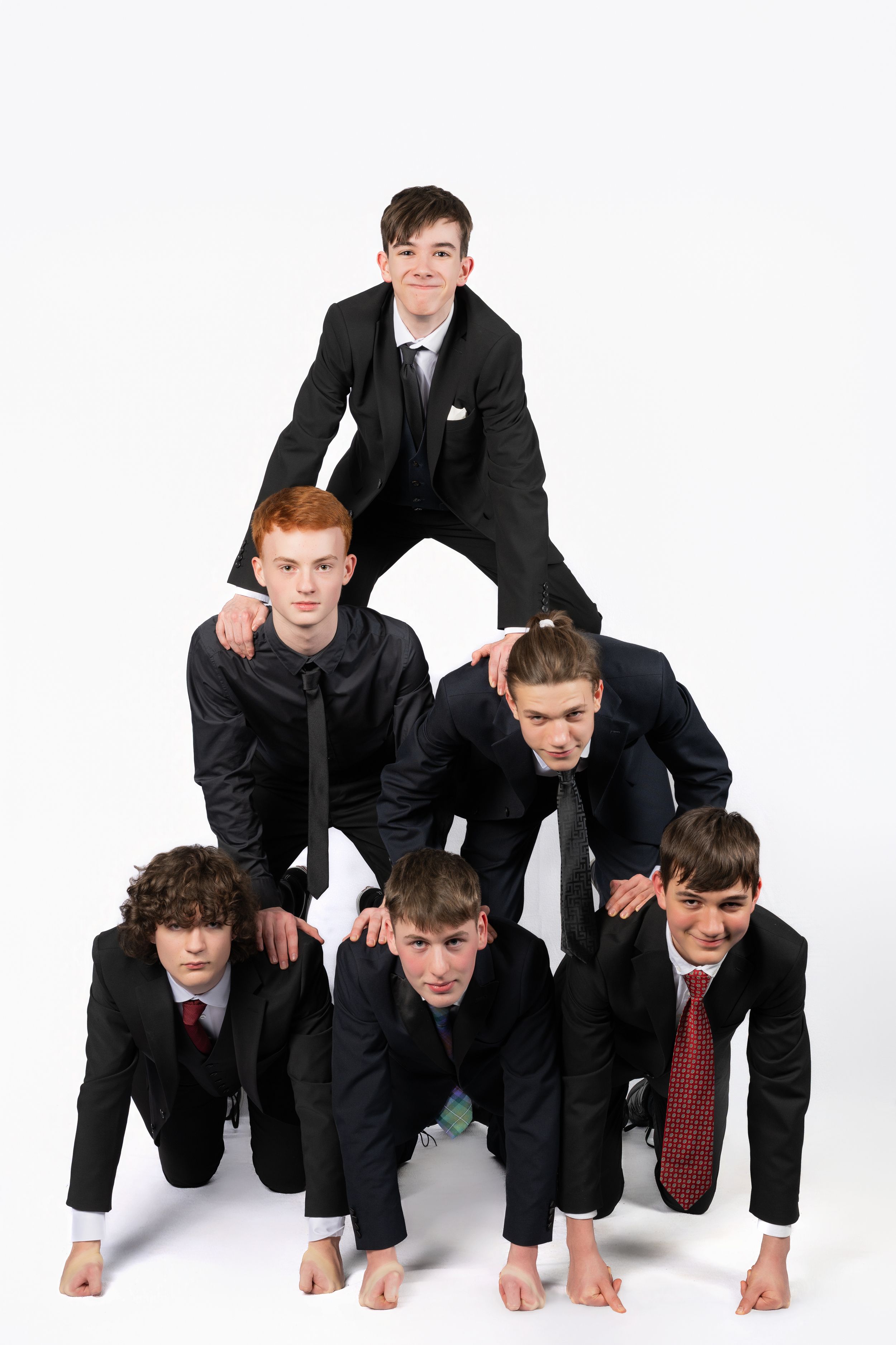 Six teenage boys in suits form a human pyramid against a white background.