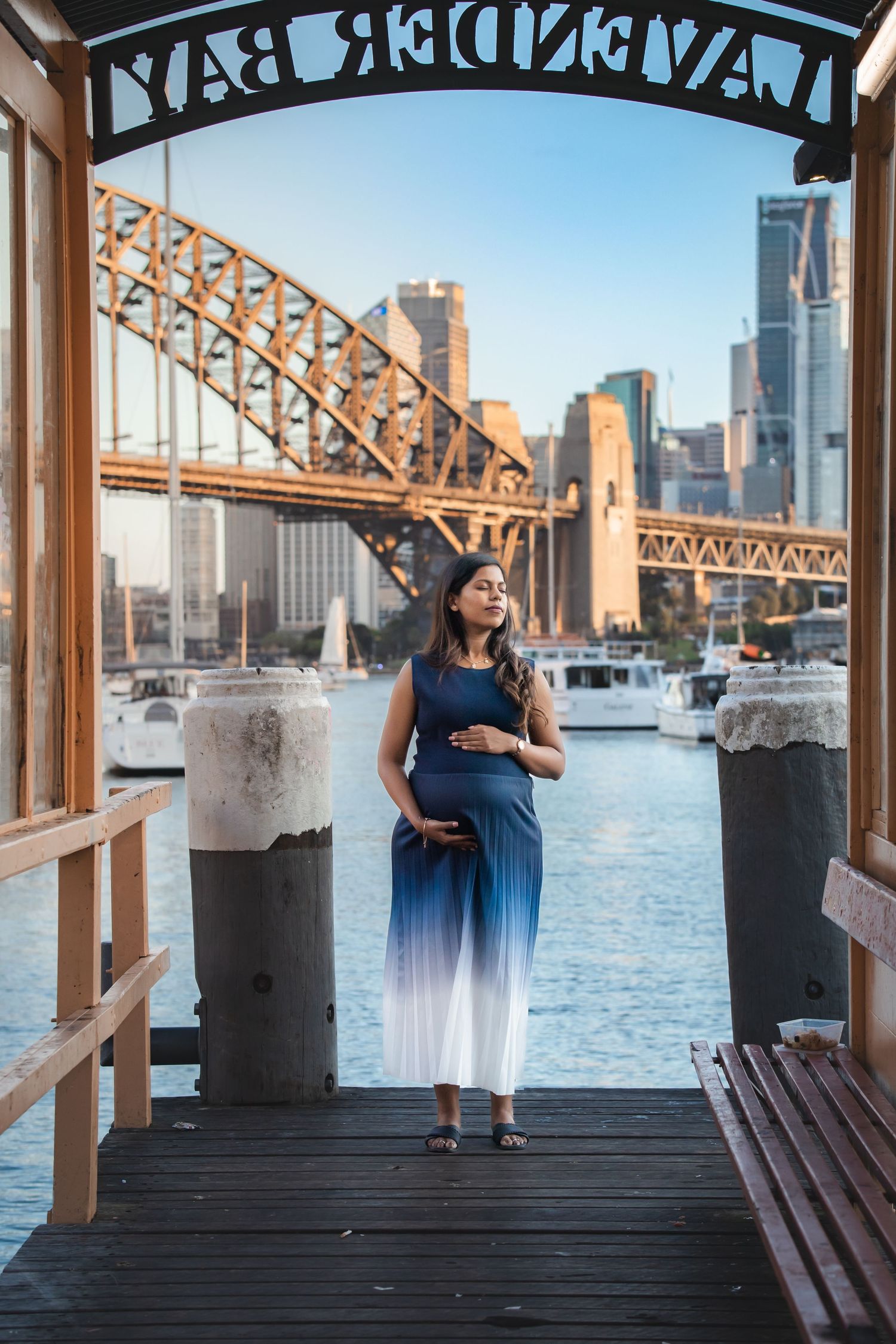 Pregnant woman in blue ombre dress poses on Lavender Bay wharf with Sydney Harbour Bridge skyline backdrop.
