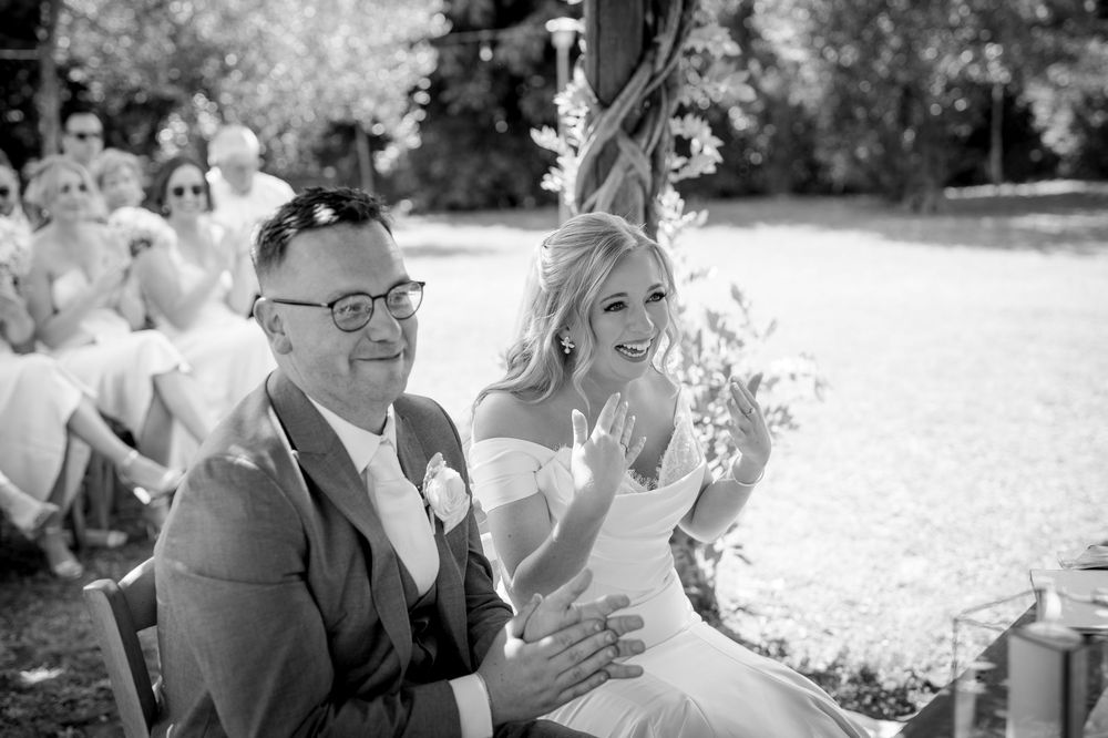 Black and white photo of smiling bride and older man seated together during outdoor wedding ceremony.