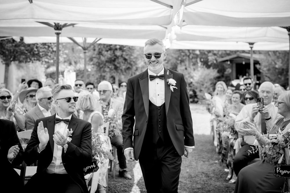 Black and white photo of groom walking down the aisle as guests applaud at outdoor wedding.