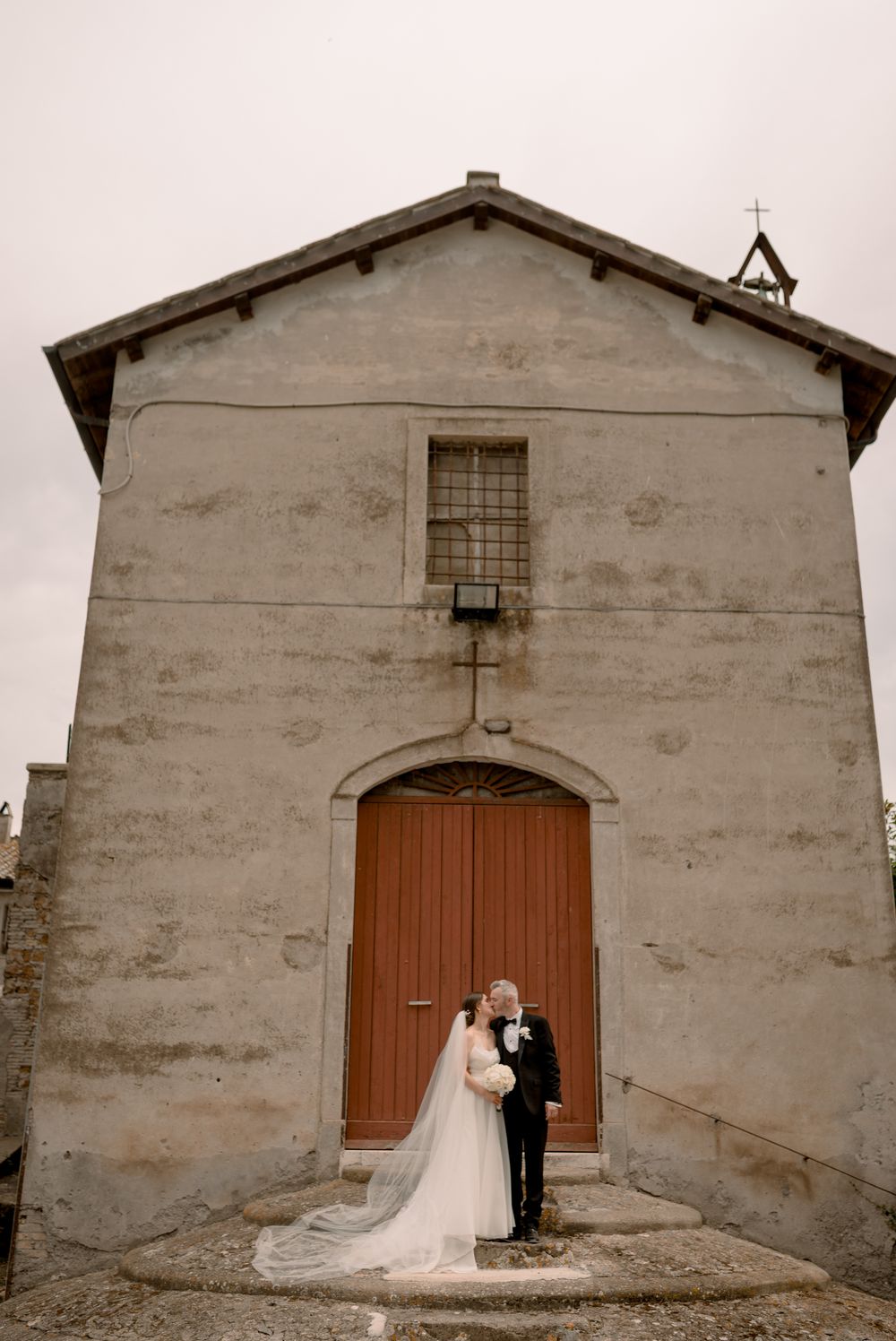 Bride and groom embracing in front of small historic stone chapel with wooden doors on wedding day.