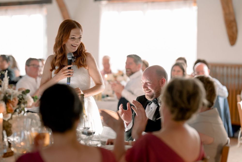 Red-haired woman giving a speech at a wedding reception while guests applaud and smile.