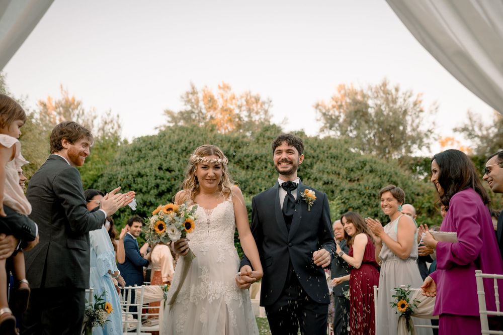 Newlyweds walk hand in hand down the aisle as guests applaud in a sunlit outdoor garden ceremony.