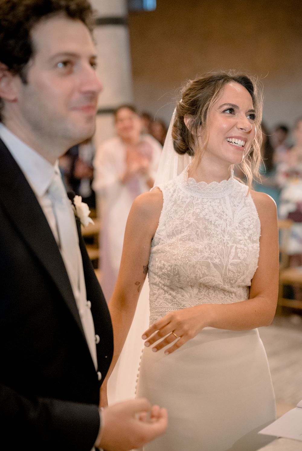 Smiling bride in beaded high-neck wedding dress stands beside groom during church wedding ceremony.