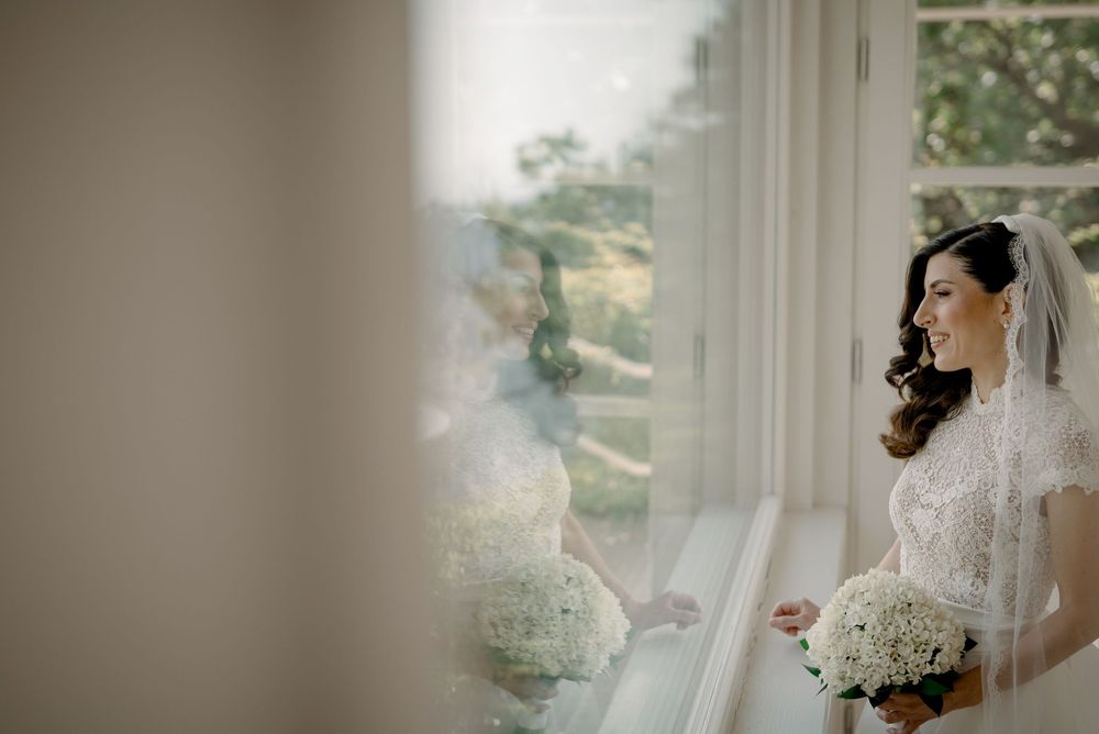 A beautiful bride in a lace veil holds a white bouquet while gazing out a window on her wedding day.