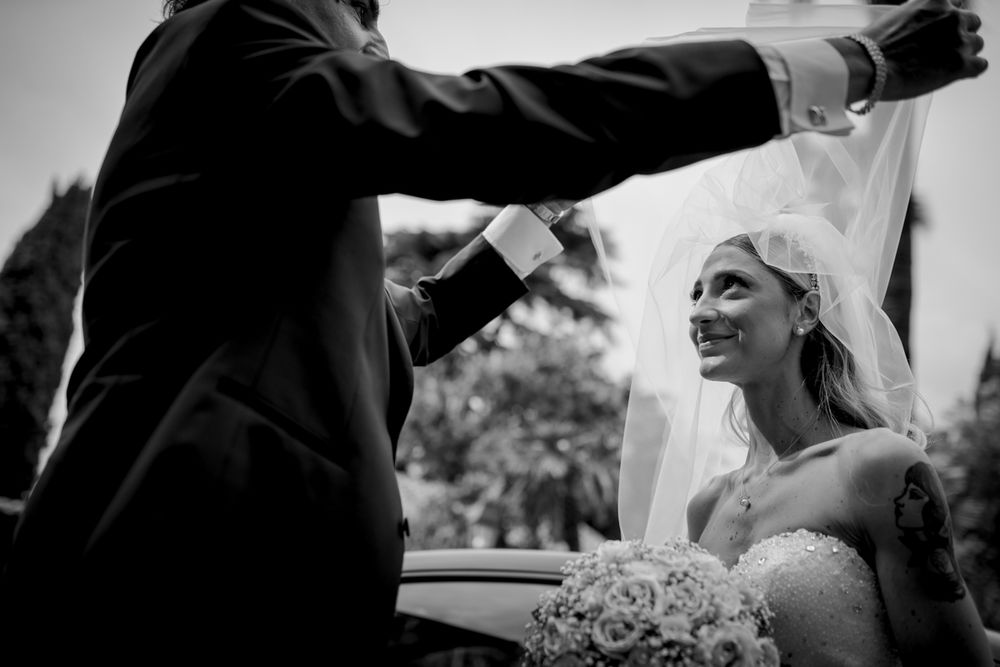 Groom lifting bride's veil outdoors as she smiles joyfully in black and white wedding photo.