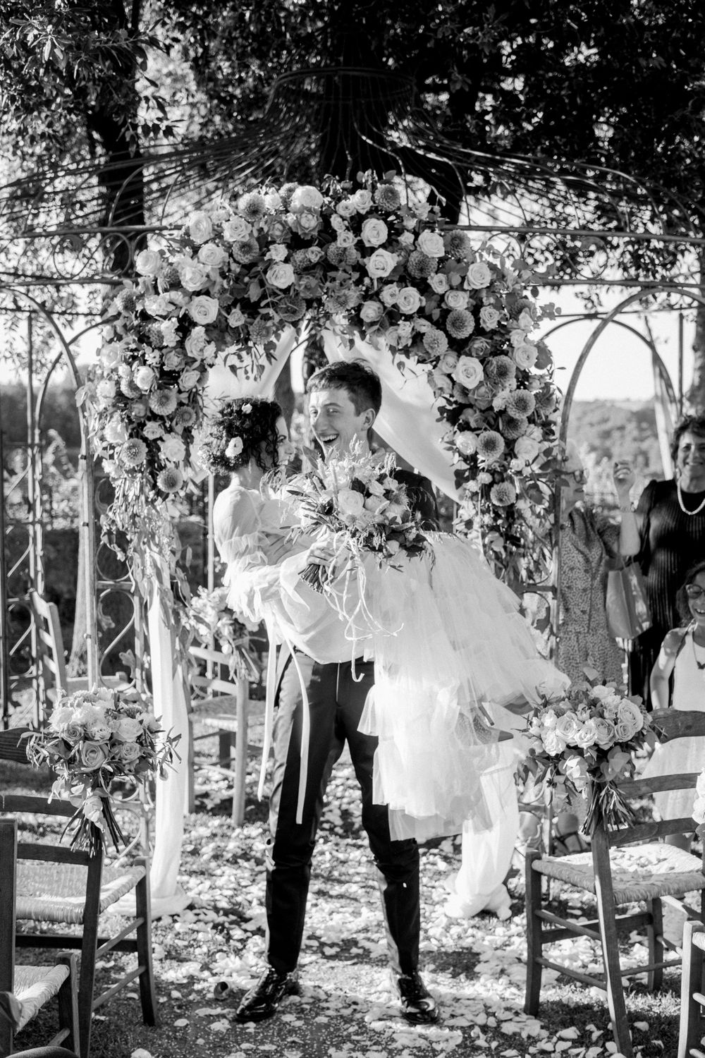 Black and white photo of groom lifting bride under a floral arch after their outdoor wedding ceremony.
