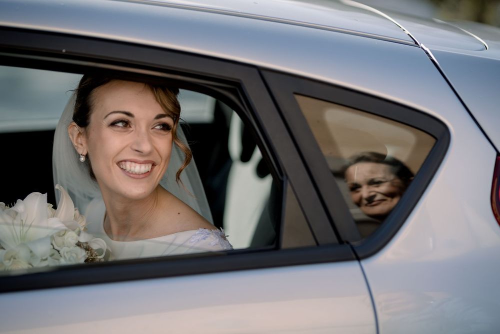 Smiling bride in veil looks out car window with reflection visible on wedding day.