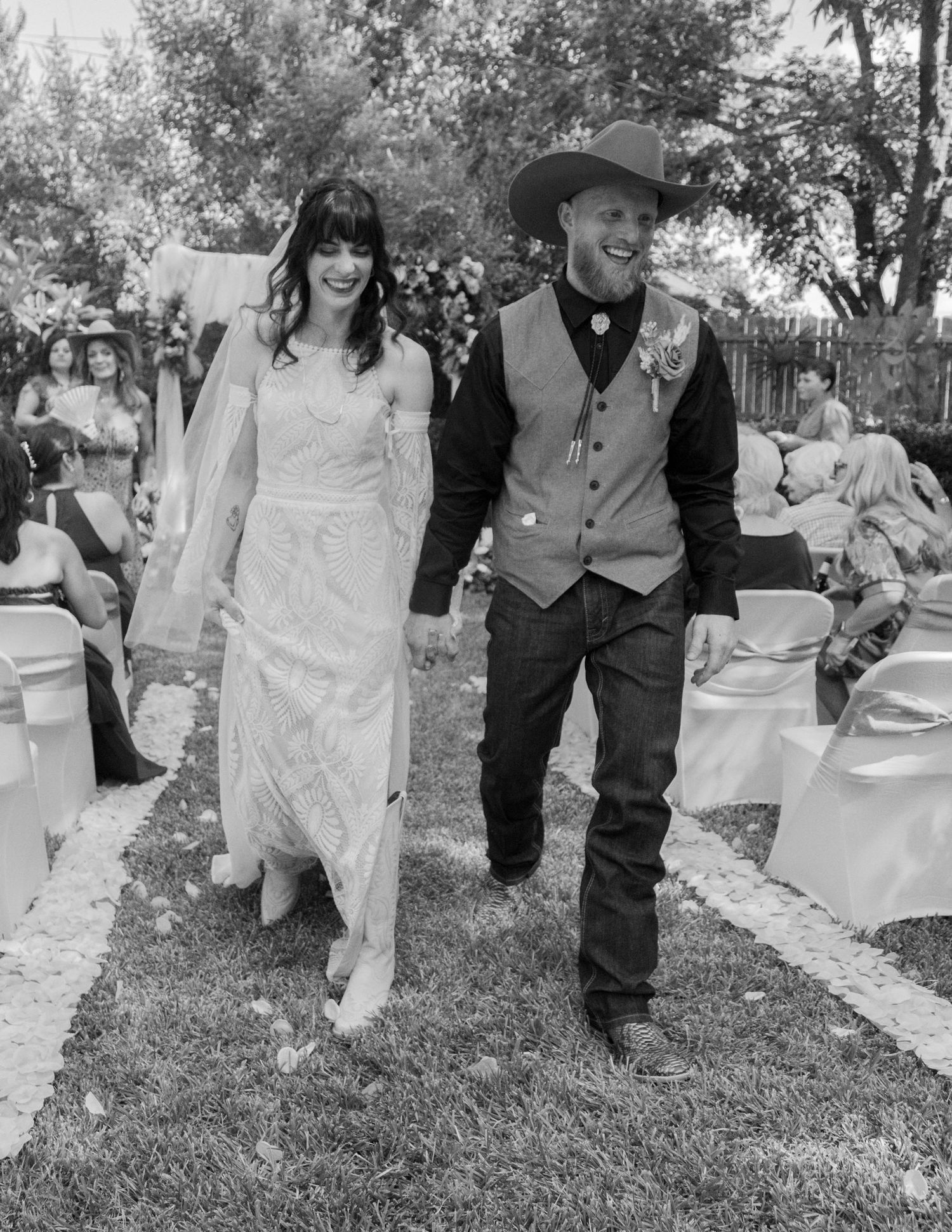 Black and white photo of a smiling bride and groom in cowboy hat walking down the aisle at an outdoor wedding ceremony.