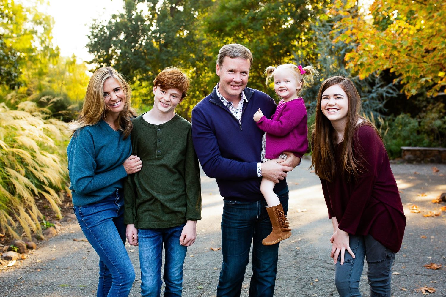 Happy family of five posing outdoors in autumn, smiling together on a garden path surrounded by colorful fall foliage.