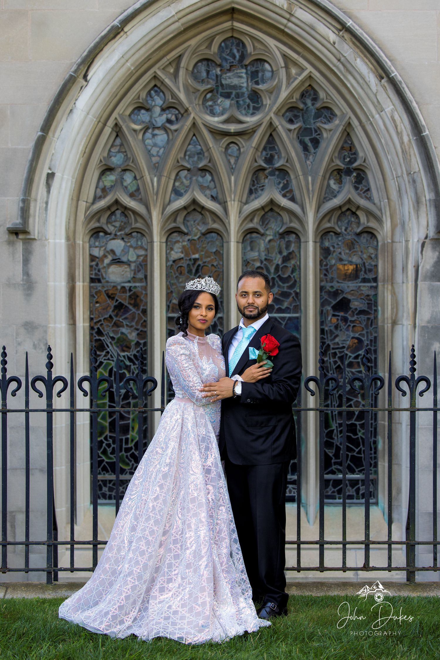 Elegant couple in wedding attire posing before a Gothic church stained glass window, bride in lace gown with tiara.