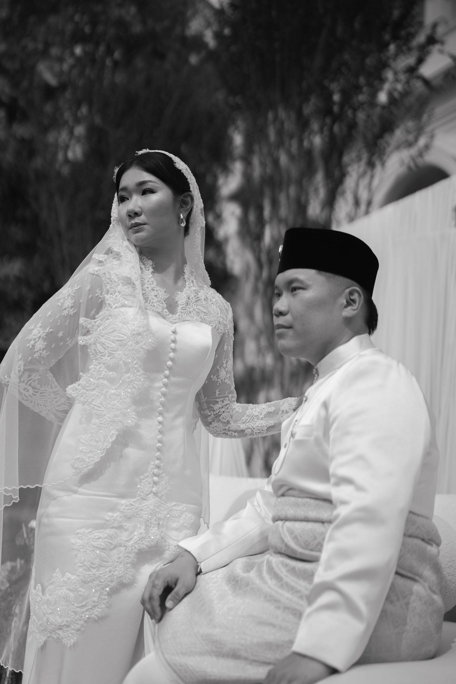Black and white photo of a Malay bride in white kebaya and veil with groom in baju melayu at a traditional wedding.