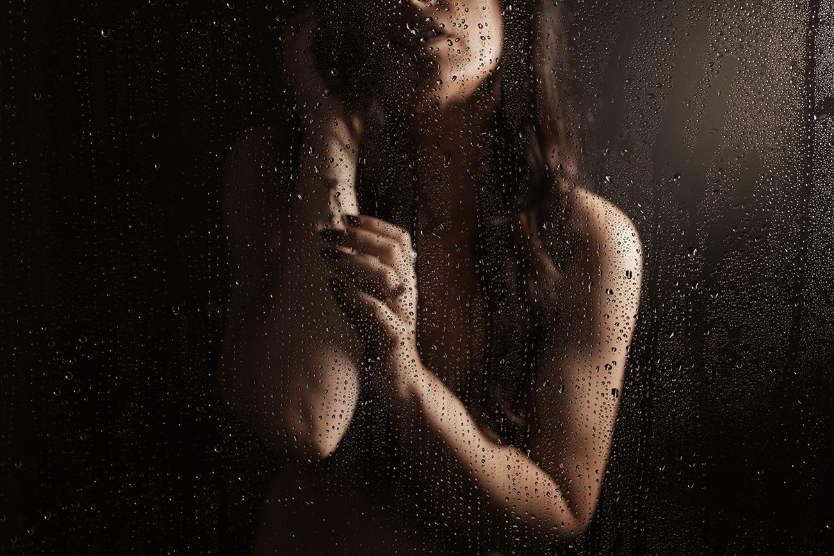 Woman standing behind rain-covered glass in dramatic moody lighting, artistic nude silhouette photography.