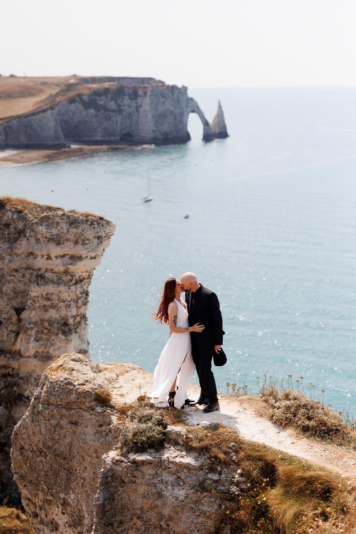 Couple embracing on cliff edge at Etretat, France, with iconic natural arch and turquoise sea backdrop.