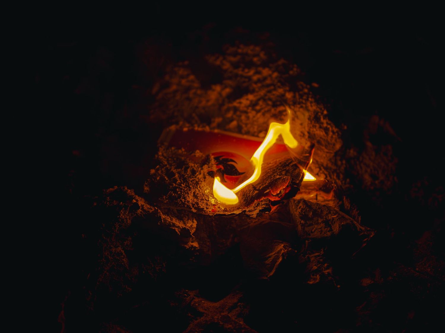 Dramatic product photograph of a collectible card burning in the dark, bright flames curling over its surface amid glowing embers and ash.
